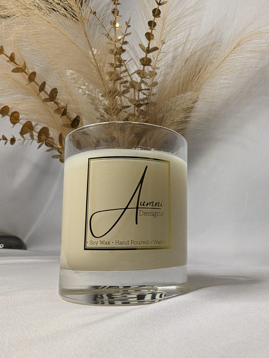 A white candle labeled 'Aumni' in a clear glass container, displayed alongside dried flowers.