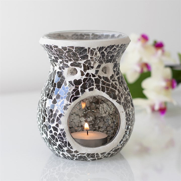 Mosaic-patterned candle holder with a lit candle on a white surface with flowers in the background